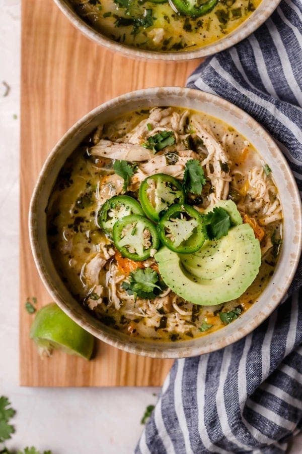 Bowl of white chicken chili with jalapeño and avocado garnish, resting on a wooden cutting board with a striped napkin surrounding it