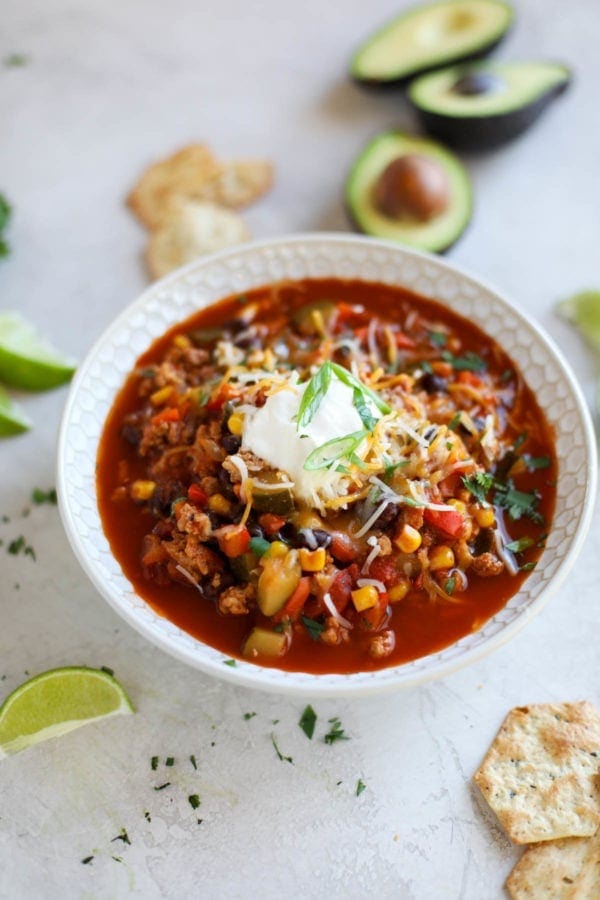 White bowl filled with veggie-loaded turkey chili topped with sour cream and cheese. Avocado, lime and crackers surround it.